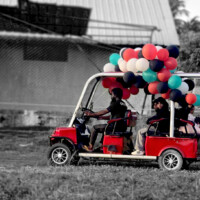 ގޮލްފް ކާޓް ރައިޑް man driving a red golf cart decorated with balloons giving rides to kids in a field